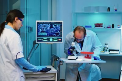Scientist researcher using microscope in laboratory while nurse taking notes on computer working overtime. Medical healthcare technology and pharmaceutical research and development concept.